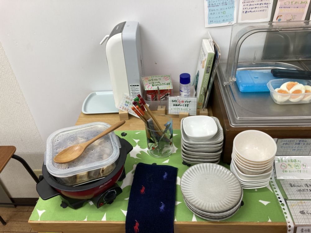 「写真の説明」この画像には、食べ物が並べられたカウンターが写っています。左側には、ゆで卵が入った透明な容器と、青い蓋の容器があります。右側には、赤い漬物のようなものが入ったガラスの容器があります。カウンターの前には、白い小皿やボウルが積み重ねられています。右側には、スナック菓子やお菓子が入ったトレイが置かれています。背景には、いくつかの手書きのメニューや案内が掲示されています。