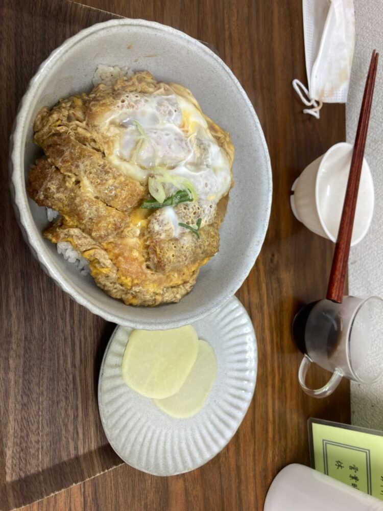 「写真の説明」写真には、カツ丼が写っています。白い陶器の丼にご飯が盛られ、その上にカツと卵が乗っています。卵は半熟で、ネギが少しトッピングされています。丼の横には、小皿に薄切りのたくあんが2枚置かれています。さらに、テーブルには箸と箸置き、そして小さなソースの入った容器が見えます。背景には、白い紙ナプキンとマスクが置かれています。