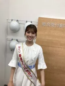 「写真の説明」Be My AI　白いワンピースを着た若い女性が笑顔で立っています。肩から「宝くじ 幸運アンバサダー」と書かれたたすきをかけています。