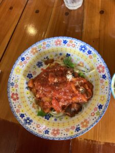「写真の説明」Be My AI - 花柄のカラフルな皿に盛り付けられたトマトソースパスタ。中央に赤いトマトソースがたっぷりかかっており、パスタの上には野菜やチーズのようなものも見えます。皿の周りには黄色や青、赤の花模様が描かれています。背景は木製のテーブルで、奥にはコップが少し写っています。