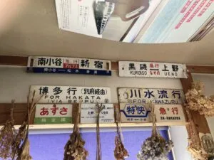 「写真の説明」Be My AI - 鉄道の行先表示板やサボ（行先標）が壁に複数飾られている様子です。上部には「南小谷 新宿」「黒磯 上野」などの表示板があり、それぞれ急行や普通列車の種別も記載されています。下の段には「博多行（別府経由）FOR HAKATA (VIA BEPPU)」「川水流行 FOR KAWAZURU」などの表示板があり、「あずさ」「特快」などの種別や指定席の案内も見えます。表示板の下にはドライフラワーが吊るされており、窓には青いカーテンがかかっています。天井近くには鉄道雑誌のページがディスプレイのように貼られています。全体的に鉄道資料館やカフェのような雰囲気です。