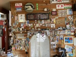 「写真の説明」Be My AI - 木の壁一面に貼られた鉄道グッズやプレート、写真、キーホルダーなどのコレクション。中央には「ED76 17」と書かれた大きな機関車のナンバープレートがあり、その周囲には「かいもん」「準急」「指定席」などの鉄道標識や行先表示板、切符、記念バッジ、名札、鉄道関連の小物がびっしりと飾られています。壁の下部にはカーテン付きのアーチ型の出入り口があり、その周りにもたくさんのグッズが吊るされています。全体的に鉄道ファンの趣味部屋やカフェの一角のような雰囲気です。