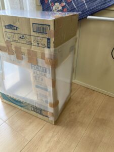 「写真の説明」Be My AI　段ボールと透明なプラスチック板で作られた箱。床の上に、段ボール箱と透明なプラスチック板を組み合わせて作られた箱が置かれています。箱の上部と下部は段ボールで、側面や正面の一部は透明なプラスチック板になっています。茶色いガムテープでしっかりと固定されています。箱の中は空で、外から中身が見える構造です。