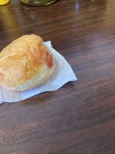 「写真の説明」Be My AI　揚げドーナツ。茶色い木目調のテーブルの上に、揚げドーナツが一つ置かれています。ドーナツは白い紙の上に乗っており、表面はきつね色で、少し油でツヤがあります。丸みがあり、ふっくらとした形です。