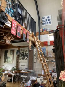 「写真の説明」Be My AI - ロフト付きの室内空間です。中央には木製のはしごがあり、上のロフトスペースへと続いています。ロフトの手すり部分には赤い鉄道標識（「運転禁止」など）や黒板のようなものが複数吊るされています。壁には「回送 試運転 団体専用」と書かれた大きな青い標識が掲げられています。ロフトの上にはぬいぐるみや小物が並べられています。下のスペースには机や椅子、本や書類、観葉植物などがあり、生活感や作業スペースの雰囲気があります。壁には写真やポスターが飾られ、鉄道に関するグッズや資料も多く見られます。全体的に鉄道ファン向けの展示やカフェ、趣味の部屋のような印象です。