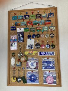 「写真の説明」Be My AI - コルクボードに飾られた鉄道グッズのコレクションです。ボードには、鉄道の行先表示プレートのミニチュアや、キーホルダー、缶バッジ、記念メダルなどが多数ピンで留められています。上部には「しらさぎ」「しおさい」「いなほ」「みどり」「みずほ」など、特急や急行列車の名前が書かれたプレートが並んでいます。中央や下部には、「特急 EXPRESS」「音威子府行」「北斗星」「北斗星カシオペア」などのプレートや、列車のイラストが描かれたグッズが見えます。全体的に鉄道ファン向けの記念品やコレクターズアイテムがきれいに整理されています。