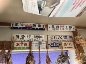 「写真の説明」Be My AI - 鉄道の行先表示板やサボ（行先標）が壁に複数飾られている様子です。上部には「南小谷 新宿」「黒磯 上野」などの表示板があり、それぞれ急行や普通列車の種別も記載されています。下の段には「博多行（別府経由）FOR HAKATA (VIA BEPPU)」「川水流行 FOR KAWAZURU」などの表示板があり、「あずさ」「特快」などの種別や指定席の案内も見えます。表示板の下にはドライフラワーが吊るされており、窓には青いカーテンがかかっています。天井近くには鉄道雑誌のページがディスプレイのように貼られています。全体的に鉄道資料館やカフェのような雰囲気です。