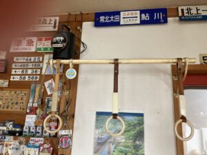 「写真の説明」Be My AI - 鉄道グッズが飾られている壁。中央には電車の吊り革が3つぶら下がっており、上部には「常北太田」「鮎川」「北上」などの駅名標が掲げられています。左側には「準急」「フェニックス」などのプレートや、車両番号プレート、キーホルダーやバッジなどの小物が多数ディスプレイされています。壁の中央下部には、鉄道が走る風景写真のポスターも貼られています。