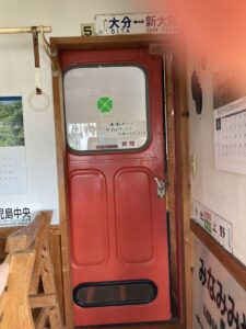 「写真の説明」Be My AI - 赤いドアとその周辺の室内の様子です。ドアの上部には「大分←→新大阪」と書かれた駅名標のような看板があり、ドア自体には窓がついています。窓には緑色のクロスのマークと、「おねがい ドアはしっかりしめてください 禁煙」と手書きのメモが貼られています。ドアの左側には吊り革があり、壁にはカレンダーや「霧島中央」などの駅名標が見えます。右側の壁にもカレンダーや「上野」などの駅名標が貼られています。