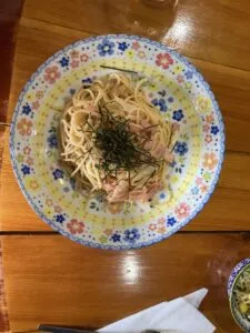 「写真の説明」Be My AI 花柄のカラフルな皿に盛り付けられたパスタ料理。パスタの上には刻み海苔と細かく切られたベーコンのような具材がのっています。木製のテーブルの上に置かれており、右下には小鉢と紙ナプキンが少し見えています。