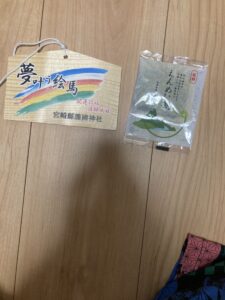 「写真の説明」Be My AI　木目の床の上に、2つのアイテムが並べて置かれています。左側は木製の絵馬で、カラフルな虹のような模様が描かれており、「夢叶う絵馬」と大きく書かれています。さらに「合格祈願　就職成就」「宮崎縣護國神社」とも記載されています。絵馬の上部には白い紐が通されています。右側は、「えんめい」と書かれたお茶が袋に入っています。